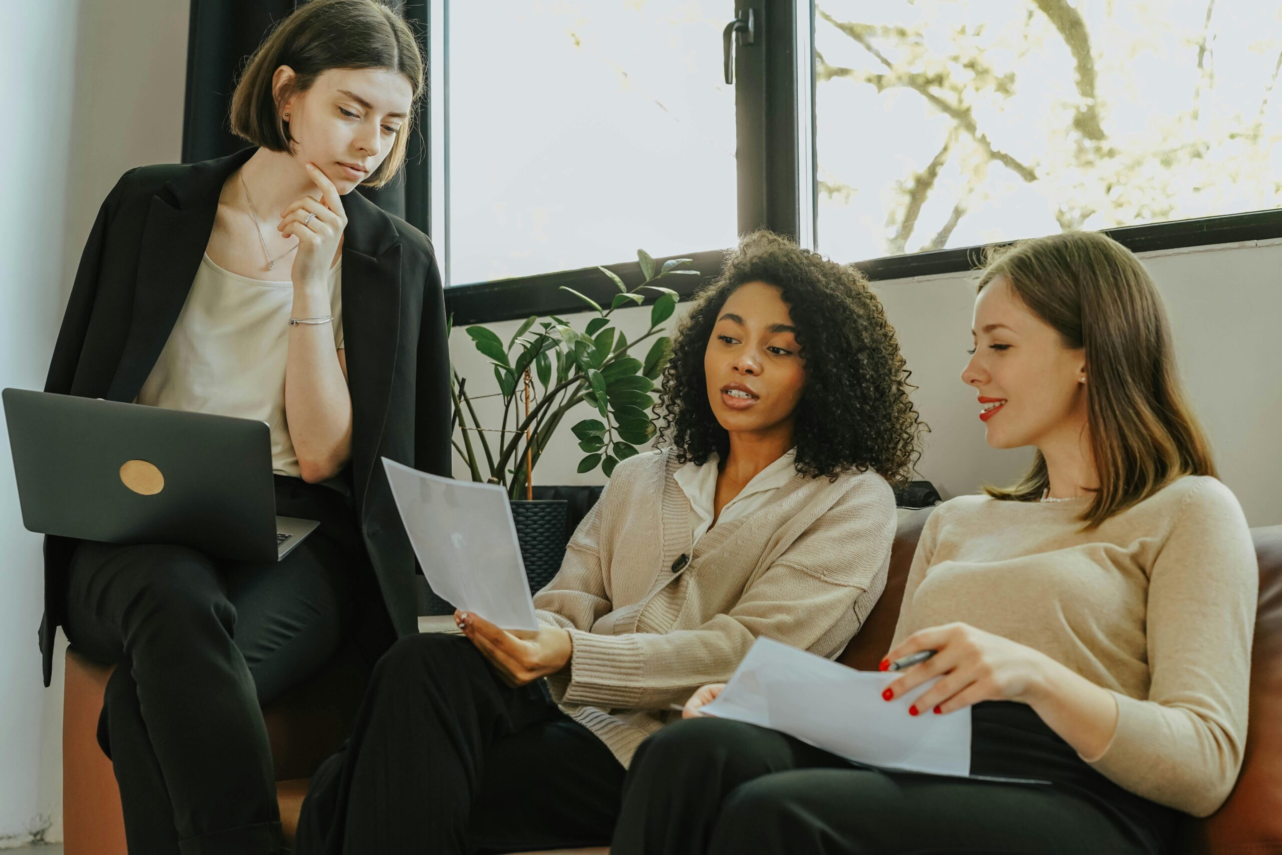NOSSOS SERVIÇOS Three professional women engaged in a collaborative meeting in a modern office setting.