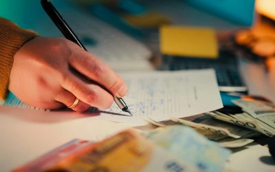Close-up of a hand writing on documents with euro notes scattered on a desk, focusing on finance and business.
